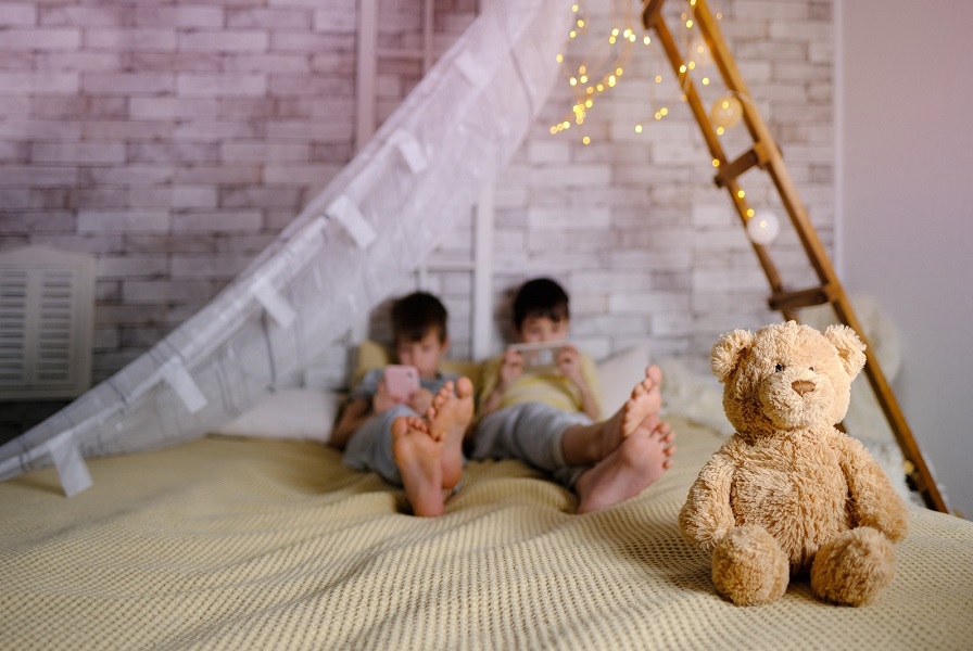 Fotografía de dos niños acostados en una cama manipulando sus dispositivos móviles e ignorando un oso de peluche. Fotografía de dos niños acostados en una cama manipulando sus dispositivos móviles e ignorando un oso de peluche.