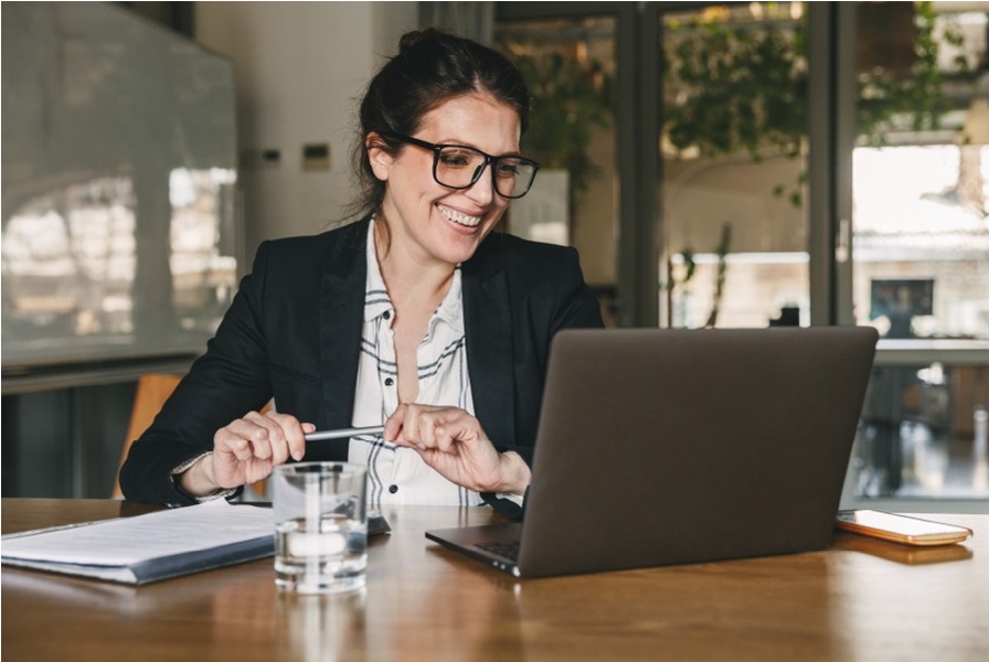 Imagen de una mujer joven que usa lentes y se muestra sonriente frente al computador portátil.