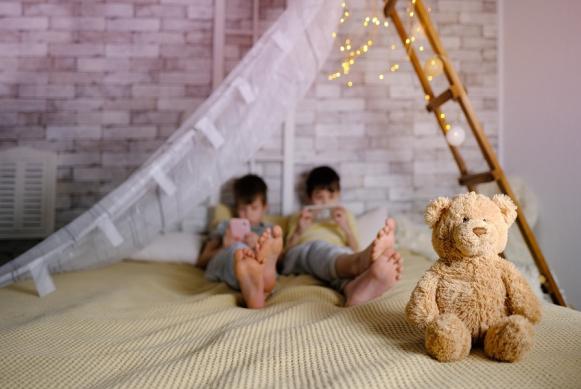Fotografía de dos niños acostados en una cama manipulando sus dispositivos móviles e ignorando un oso de peluche. Fotografía de dos niños acostados en una cama manipulando sus dispositivos móviles e ignorando un oso de peluche.